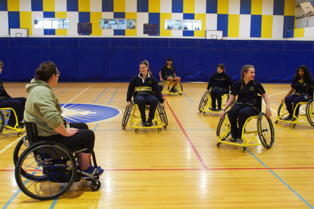 A group of students are using wheelchairs on an indoor basketball court